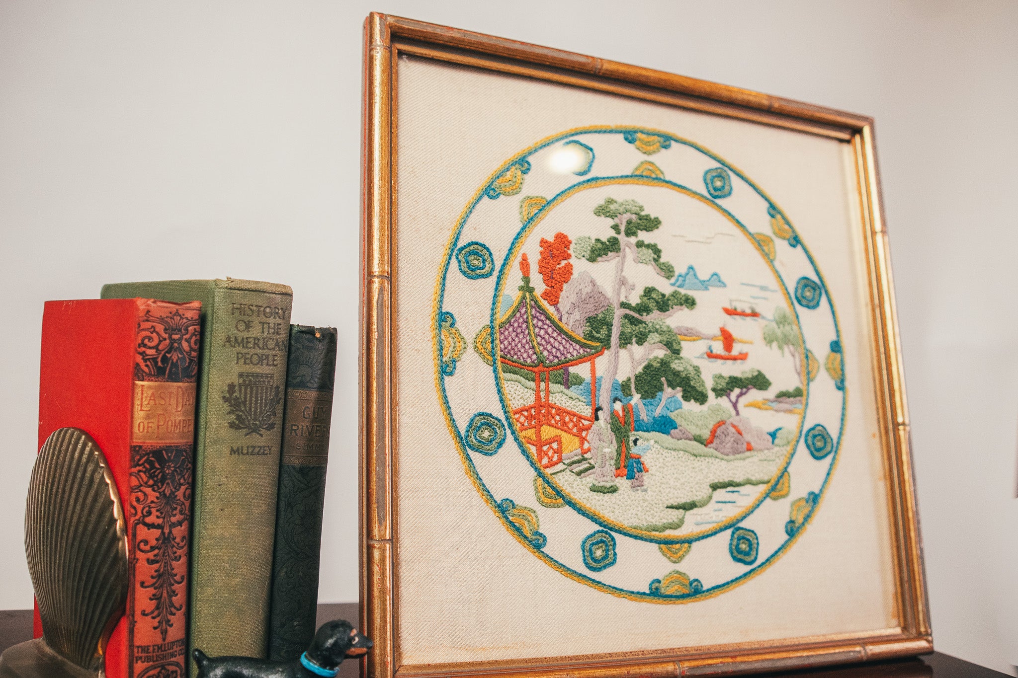 Framed embroidered artwork with a scenic design on a table with books.