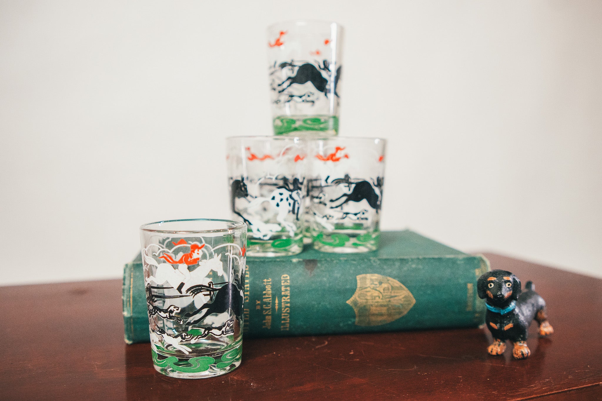 Set of glass tumblers with horse design on a wooden table with a book and small dog figurine.