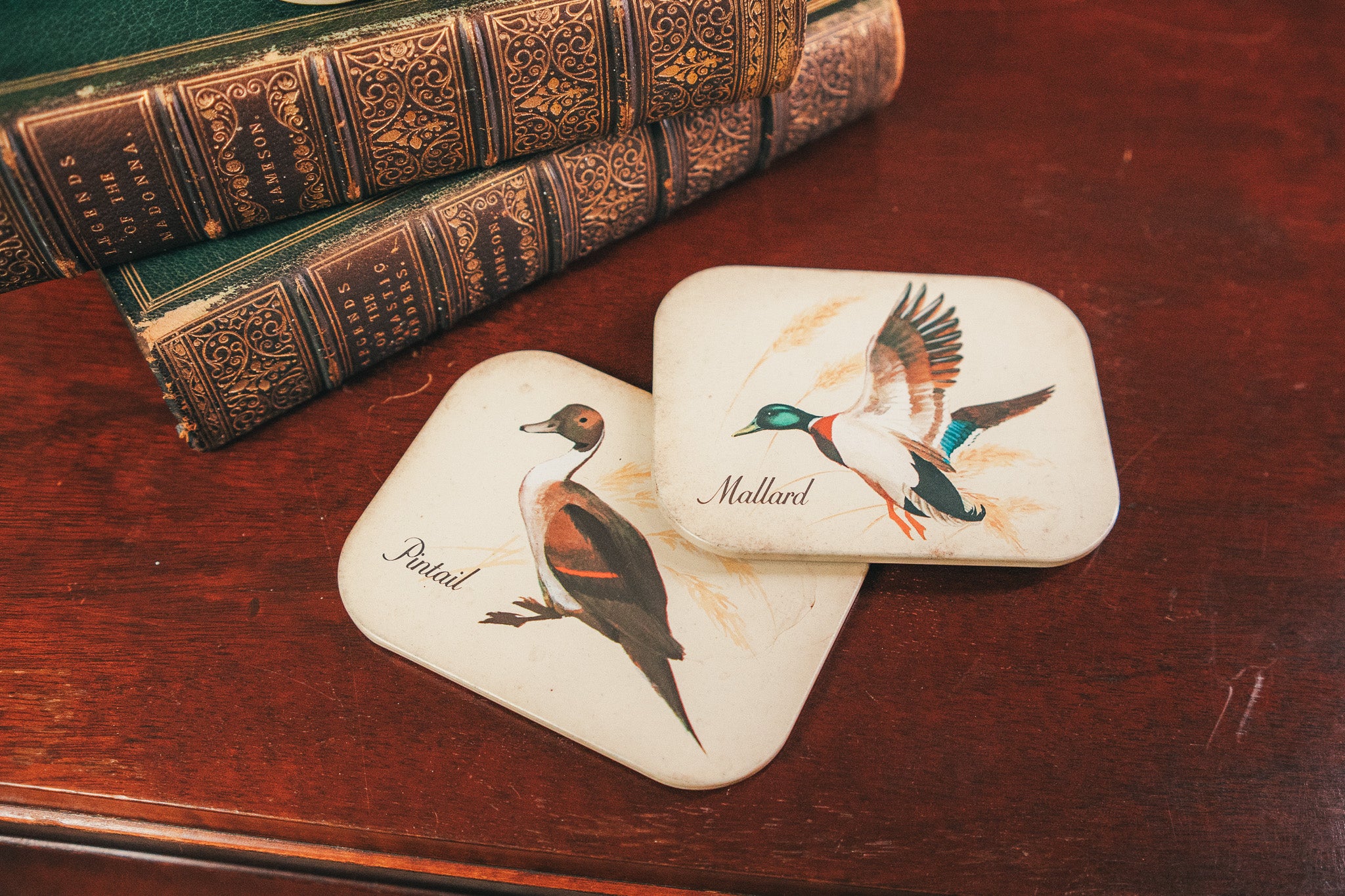 Two coasters with duck illustrations on a wooden surface with vintage books.