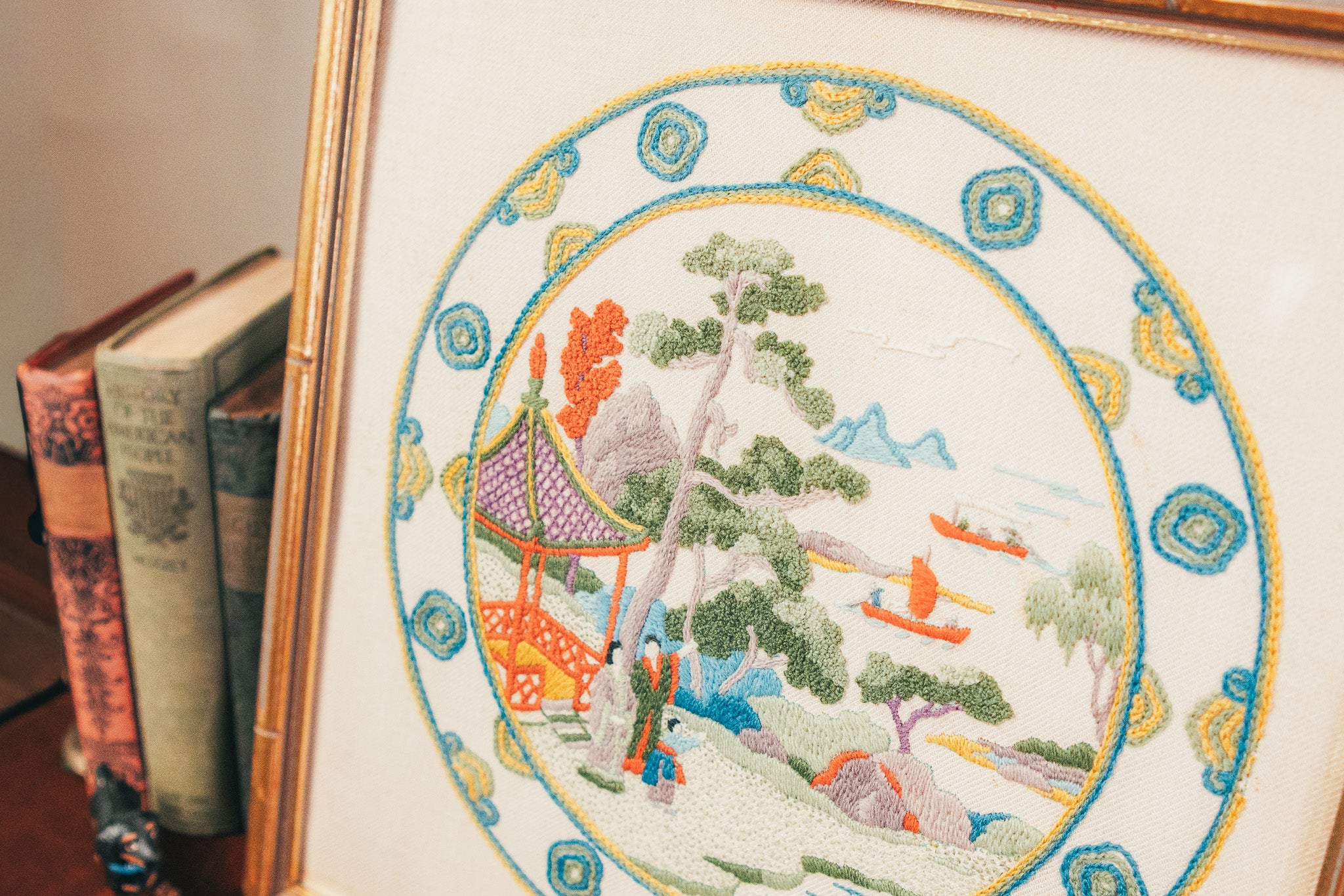 Framed embroidered artwork depicting a scenic landscape with trees and people, placed on a surface with books.